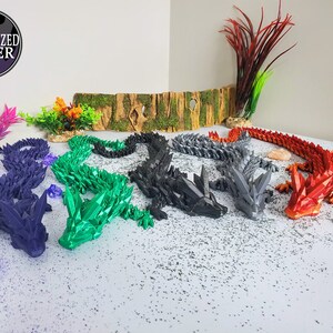 May include: A collection of articulated dragon figurines in various colors, including purple, green, black, gray, and red. The dragons are posed on a white surface with decorative elements. Each dragon has a crystal-like design.