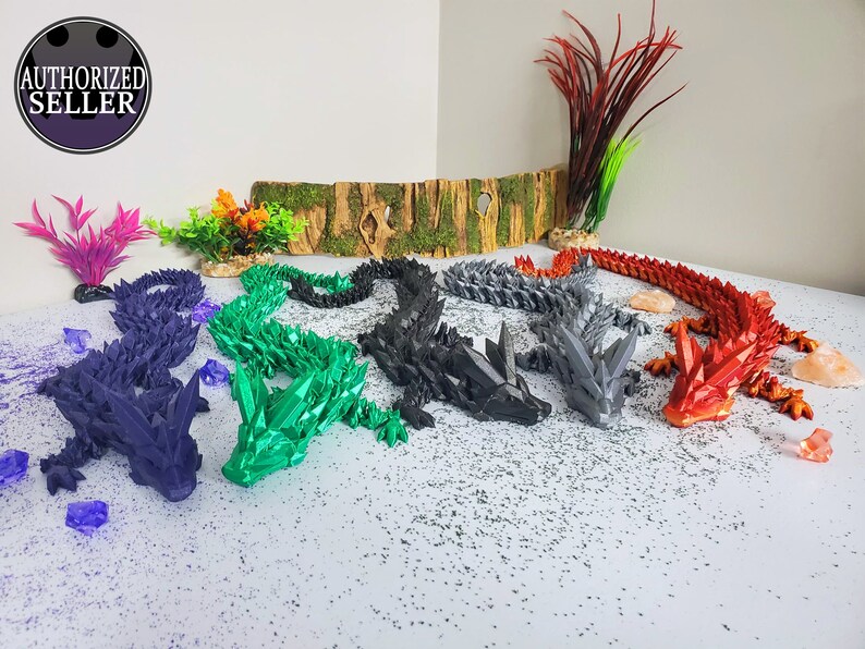 May include: A collection of articulated dragon figurines in various colors, including purple, green, black, gray, and red. The dragons are posed on a white surface with decorative elements. Each dragon has a crystal-like design.