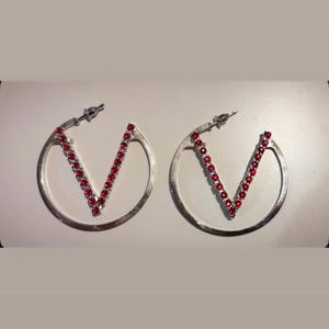 Puede incluir: Un par de pendientes de aro plateados con una barra interior en forma de V incrustada con pequeñas piedras redondas de color rojo vibrante. Los pendientes tienen un cierre de presión y un acabado metálico pulido, creando un diseño circular.
