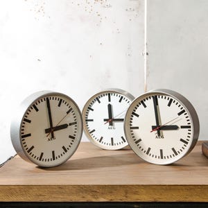 May include: Three round analogue clocks with silver-toned metal frames and off-white faces. Each clock features bold black hour and minute hands, a thin red second hand, black tick marks for the hours and minutes, and the black text AEG.