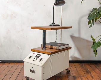 May include: An industrial-style side table with a cream-colored metal base and a wooden top. The table features a black desk lamp and a book. The table has wheels for easy movement. The table is a unique piece of furniture.