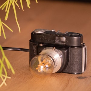 Peut inclure: Un appareil photo vintage noir et argent avec une ampoule attachée à l'objectif. L'appareil photo porte l'inscription "Beinette" sur le devant.