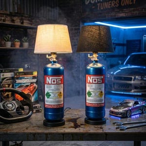 May include: A white ceramic mug with a black handle and the text "But First, Coffee" in a bold, black font. The mug is set against a blurred background, suggesting a cozy setting.