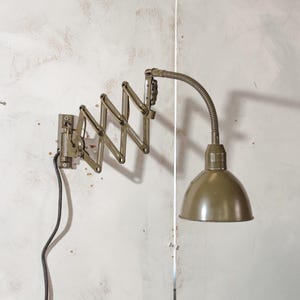 Può includere: Una lampada da parete in stile industriale verde oliva. La lampada presenta un braccio a fisarmonica e un collo di cigno flessibile che conduce a un paralume a cupola. Un cavo di alimentazione nero si estende dalla base, montato su una parete bianca strutturata.
