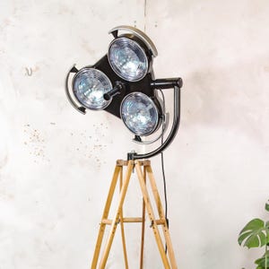 May include: A black and silver industrial-style floor lamp with three circular lights. The lights are mounted on a black frame and supported by a wooden tripod base. The lamp has a vintage aesthetic.