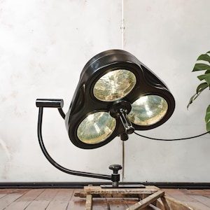 May include: Black vintage surgical lamp with three circular light fixtures. The lamp is mounted on a curved arm and a wooden base. The light fixtures have a reflective surface.