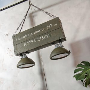 Può includere: Lampada a sospensione in stile industriale. Presenta una scatola di legno riciclata verniciata verde oliva, con testo bianco a stencil. Due paralumi in metallo verde oliva con vetro trasparente sono attaccati sotto. Sospesa tramite una catena.