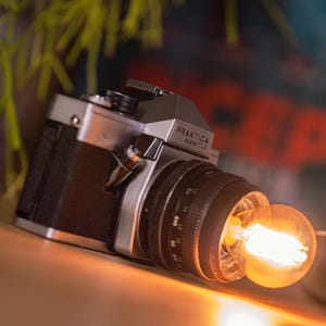 Könnte beinhalten: Eine silber-schwarze Praktica super TL 2 Filmkamera mit schwarzem Objektiv. Eine leuchtende, warm getönte Edison-Glühbirne ist direkt vor dem Objektiv positioniert und wirft ein helles orangefarbenes Licht auf das Kameragehäuse und die Oberfläche.