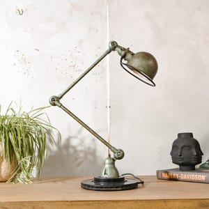 Peut inclure: Lampe de bureau de style industriel avec une finition vert vieilli. Elle possède un bras réglable et un abat-jour rond avec un bord noir. La base est un composant circulaire de couleur foncée. La lampe est posée sur une surface en bois.