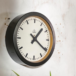 Può includere: Un orologio da parete rotondo con cornice nera, quadrante bianco e lancette nere. L'orologio ha un anello interno dorato e segni neri per le ore e i minuti. L'orologio è montato su una parete bianca.