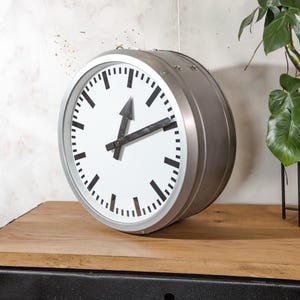 May include: A large, round, silver-colored wall clock with a white face. The clock features black hour and minute markers, and black hands. The clock is resting on a wooden surface.