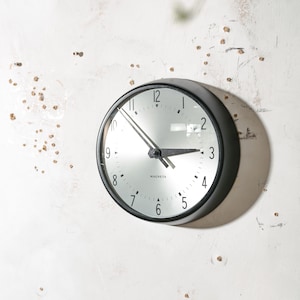 May include: A round wall clock with a black frame and a reflective silver face. The clock features black Arabic numerals from 1 to 12, black hour and minute hands, and the word MAGNETA printed in black text below the center of the clock face.