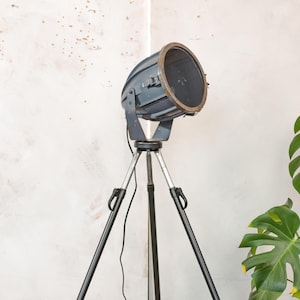 May include: A vintage-style, industrial floor lamp with a dark gray spotlight head and a tripod base. The spotlight has a circular opening with a bronze-colored rim. The tripod legs are black and silver, creating a striking contrast. The lamp is designed for interior lighting.