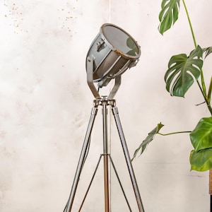 May include: A silver tripod floor lamp with a large, round, gray metal spotlight. The lamp is angled slightly to the right and the spotlight is facing forward.