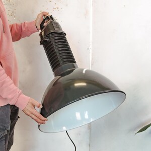 Może przedstawiać: Duża lampa wisząca w stylu industrialnym z czarnym, kopulastym kloszem i białym wnętrzem. Lampa ma żebrowaną, elastyczną szyję i uchwyt u góry. Lampa jest trzymana przez osobę.