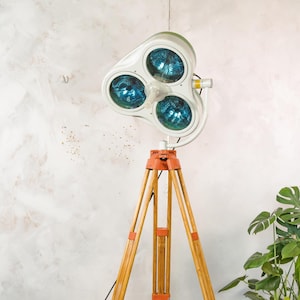 May include: A vintage medical examination lamp with three blue-tinted lights. The white lamp head is mounted on a wooden tripod stand with orange accents. The lamp is a unique piece of industrial decor.