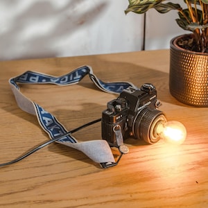 Könnte beinhalten: Eine schwarze Vintage-Kamera mit einem blau-weißen Band wurde in eine Tischlampe mit einer Glühbirne im Objektiv umgewandelt. Die Kamera steht auf einer Holzoberfläche.