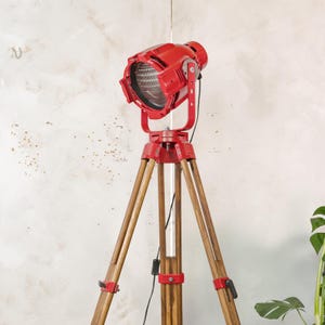 May include: A bright red spotlight lamp mounted on a tall, three-legged wooden tripod stand. The lamp features a circular glass lens, a metal housing with adjustable side hinges, and a black power cord with an inline switch attached to the frame.