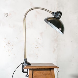 Puede incluir: Una lámpara de escritorio negra con un brazo flexible y una base de abrazadera. La lámpara tiene una pantalla de metal negro y un brazo de metal dorado.