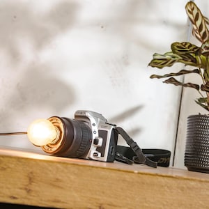 Puede incluir: Una cámara Minolta Dynax 300si plateada con una lente negra y una correa de cámara negra. La cámara está sobre una superficie de madera con una bombilla en la lente.