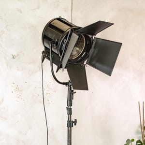 May include: A black studio spotlight with adjustable barn doors, mounted on a black stand. The spotlight has a cylindrical body and a wire mesh covering the light source. The stand is adjustable and has a series of locking mechanisms.