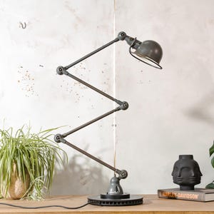 Puede incluir: Una lámpara de escritorio articulada gris con una pantalla redondeada y un diseño de brazo en zigzag. La lámpara es de metal y tiene una base circular. La lámpara está sobre una superficie de madera. Una planta en maceta y un jarrón decorativo están en el fondo.