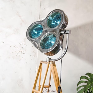 May include: A vintage-style floor lamp with three circular light fixtures. The lamp features a brushed metal finish and blue-tinted glass. It is mounted on a wooden tripod base with a black power cord and switch. The lamp is designed for interior lighting.