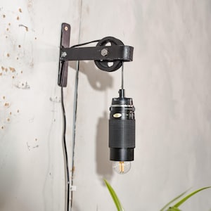Könnte beinhalten: Eine einzigartige, schwarze Wandlampe aus einem wiederverwendeten Kameraobjektiv. Die Lampe verfügt über ein Flaschenzugsystem und eine sichtbare Glühbirne. Die Leuchte ist an einer dunkelbraunen Holzhalterung befestigt und hat ein schwarzes Netzkabel.