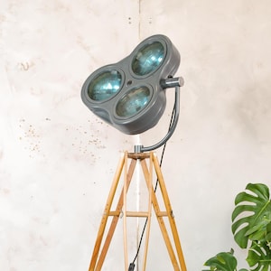 May include: A gray, three-bulb spotlight lamp on a wooden tripod base. The lamp has a curved arm and a power cord. The wooden tripod has a natural finish. The background is a neutral color with a plant in the corner.