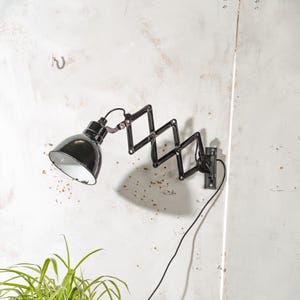 Può includere: Lampada da parete nera con un braccio estensibile a fisarmonica e un paralume nero. La lampada è montata su una parete bianca e ha un cavo di alimentazione nero. Il design della lampada è industriale e vintage.