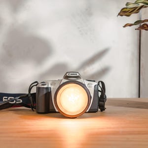 Könnte beinhalten: Eine silberne Canon EOS 500 Kamera mit einem schwarzen Objektiv und einem schwarzen Gurt. Die Kamera befindet sich auf einer Holzoberfläche.