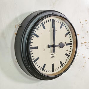 Può includere: Un orologio rotondo con cornice nera e quadrante color crema. L'orologio ha lancette e segni neri. L'orologio è montato su un muro bianco. L'orologio segna circa le 3.