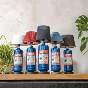 Puede incluir: Cinco lámparas azules con forma de botella NOS con pantallas de diferentes colores: blanco, rojo, negro, azul y negro. Cada lámpara tiene una etiqueta con el texto "NOS" e información de seguridad. Las lámparas se exhiben en una estantería de madera.