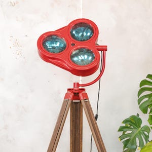 May include: A red spotlight with three blue-tinted lenses sits atop a wooden tripod. The lamp's red frame contrasts with the brown wooden legs. A black power cord extends from the lamp.