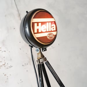 Puede incluir: Lámpara de foco Hella vintage en negro y marrón. La lámpara tiene una cara redonda, roja y blanca con la palabra "Hella" en blanco. Está montada sobre una base de trípode con patas negras y detalles metálicos. Diseño retro.