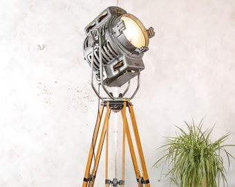 Peut inclure: Un projecteur industriel en métal argenté monté sur un grand trépied en bois à trois pieds. Le projecteur présente une lentille circulaire émettant une lumière chaude et vive, avec des fentes de ventilation et des boutons de réglage.