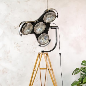 May include: A black and silver spotlight lamp with five lights, mounted on a wooden tripod base. The lamp has a modern design with a black frame and silver accents. The tripod is made of light-colored wood.
