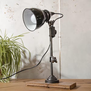 Puede incluir: Lámpara de escritorio de estilo industrial en blanco y negro. La lámpara tiene una pantalla de metal negro con interior blanco, un brazo flexible y una base redonda. Está montada sobre una base de madera y tiene una bombilla visible.