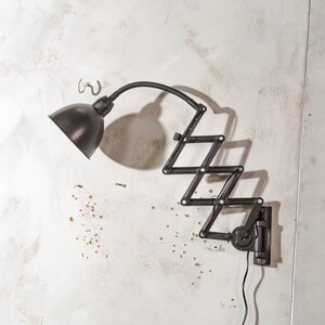 Può includere: Una lampada da parete nera in stile industriale con braccio regolabile. La lampada ha un paralume arrotondato e un braccio a fisarmonica che si estende dalla parete. La lampada è progettata per dirigere la luce.