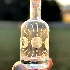 May include: A clear glass bottle with a gold lid and a moon phase design etched into the glass. The bottle is being held in a hand.