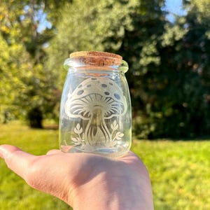 May include: A clear glass jar with a cork lid and an etched design of a mushroom with leaves. The jar is sitting on a person's hand.