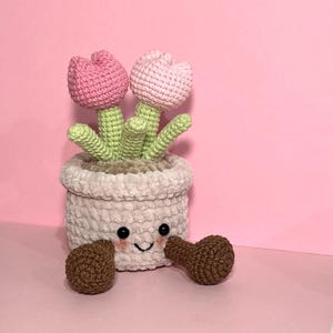 May include: A crocheted pink tulip flower pot with a smiling face and brown crocheted arms. The pot is filled with green crocheted leaves and two pink crocheted tulips.