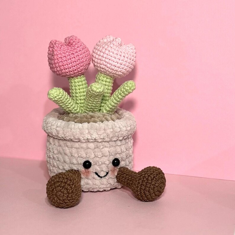 May include: A crocheted pink tulip flower pot with a smiling face and brown crocheted arms. The pot is filled with green crocheted leaves and two pink crocheted tulips.