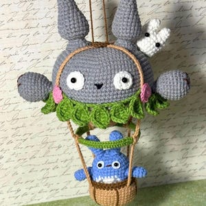 May include: A handmade crochet mobile featuring a gray character with large ears, white eyes, and a small black nose. A smaller blue character is in a brown basket suspended below. The mobile includes green leaves and pink flowers.