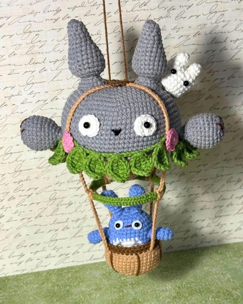 May include: A handmade crochet mobile featuring a gray character with large ears, white eyes, and a small black nose. A smaller blue character is in a brown basket suspended below. The mobile includes green leaves and pink flowers.