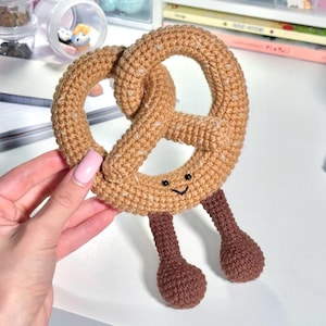 Peut inclure: Un jouet bretzel au crochet en fil marron clair avec des accents beige mouchetés. Le bretzel présente un visage noir brodé avec deux petits yeux et un sourire. Deux jambes au crochet marron foncé avec des pieds arrondis dépassent du bas.