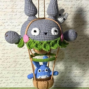 May include: A handmade crochet plush toy featuring a large gray character with a smaller blue character in a basket. The gray character has large eyes, ears, and a green collar. The blue character is in a brown basket suspended by a brown string.