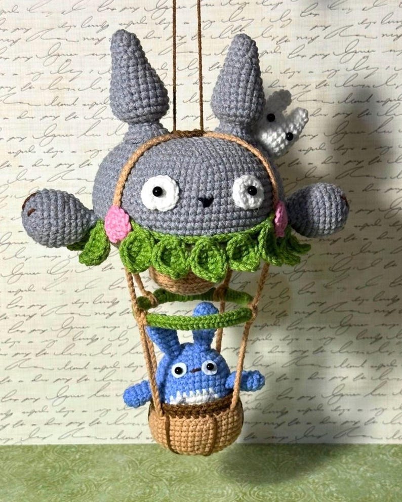 May include: A handmade crochet plush toy featuring a large gray character with a smaller blue character in a basket. The gray character has large eyes, ears, and a green collar. The blue character is in a brown basket suspended by a brown string.