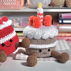 Op de afbeelding: Een gehaakte cupcake-knuffel met een beige basis, witte glazuur, oranje bessen en een kaarsje met een gele vlam. Het heeft zwarte veiligheidsoogjes en een geborduurde glimlach, met bruine armen en benen. Daarnaast staat een rode gehaakte knuffel.