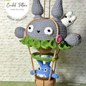 May include: A crocheted gray character with a white bunny and a blue character in a basket, suspended by strings. The gray character has large eyes and a green leafy collar. The image includes the text "Crochet Pattern" and "Digital Download".